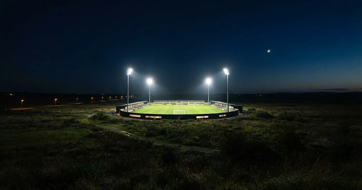 Stadion piłkarski nocą widziany z dużej odległości, jasne reflektory nad murawą i ciemne niebo wokół.