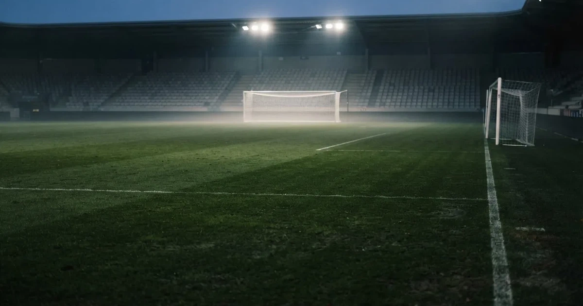 Pusta murawa stadionu miejskiego w chłodny jesienny wieczór, siatka bramki świeci w świetle reflektorów, nad boiskiem wisi mgła.